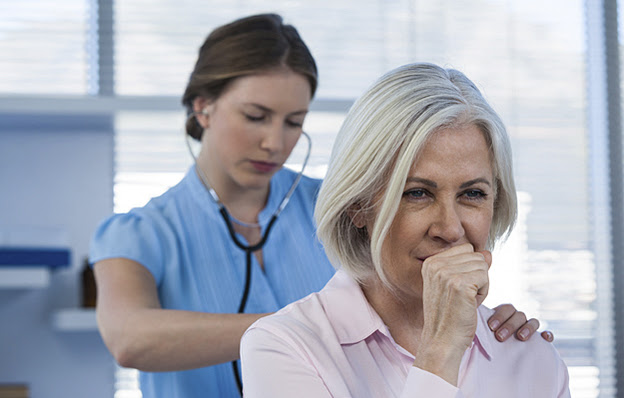 A senior woman coughing while a doctor listens with a stethoscope.