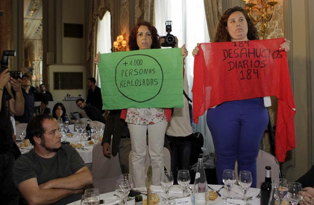 El dipuado de la CUP David Fernández observa a las dos activistas de la PAH que muestran en silencio pancartas contra los desahucios durante la intervención de Cañete.