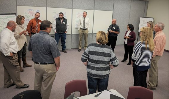 A dozen educators stand in a circle in the middle of a conference room for a facilitated discussion.