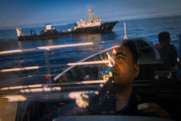 A man’s face is seen through glass on a boat against the reflection of a different boat in the South China Sea.