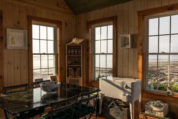 The homey interior of a cabin is decorated with a table, a vintage-style metal cooler and framed art. An island and the sea beyond can be seen through large windows.