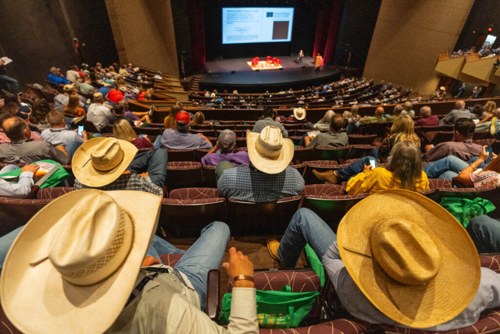 Ranchers wearing cowboy hats fill the top rows of the Rudder auditorium and a crowd fills chairs all the way down to the stage