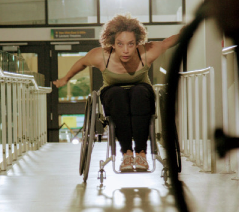An African American woman seated in a wheelchair, arms outstretched to propel herself down a ramp, faces the camera with eyes locked.