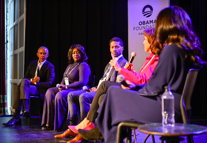 President Obama listens as Marion Gross speaks on an economic development panel in Chicago, IL, alongside Nicole Hayes, Charles Smith, and Lata Reddy. All panelists have a range of skin tones and sit on a stage with signage in the background that reads, “Obama Foundation.”