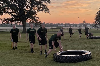 two small groups of men with shaved heads, dark green T-shirts and black shorts take turns lifting and moving large black tractor tires outdoors