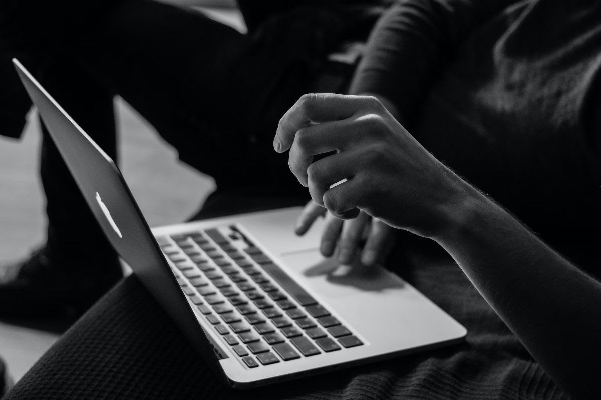 A person on a silver Apple laptop, their right hand on the mouse, their left raised in the foreground.