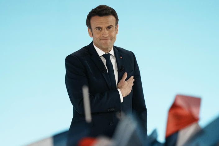 President Emmanuel Macron, wearing a rueful expression, puts his hand on his heart, with a foreground of tricolor flags.