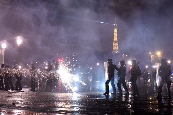 Police officers holding riot shields at night, with a bright white burst of light near the middle of the scene and the Eiffel Tower in the background.
