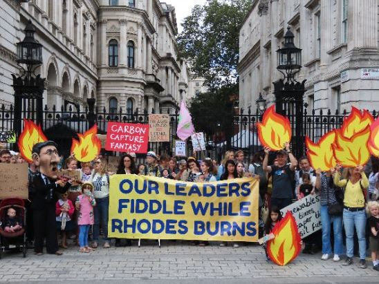 A large group of people outside Downing Street. A large banner in the centre reads Our leaders fiddle while Rhodes burns
