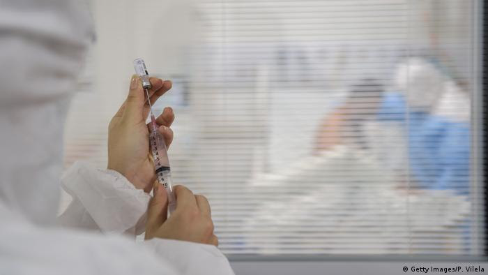 Funcionário de equipe médica prepara injeção em frente à sala de paciente em atendimento.