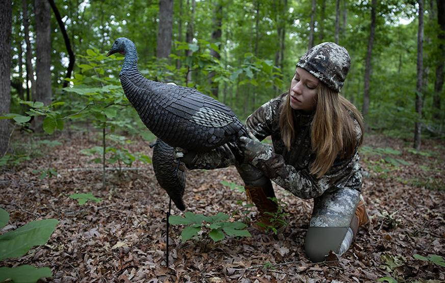 Young woman hunter wearing camouflage stands with turkey decoy in woods