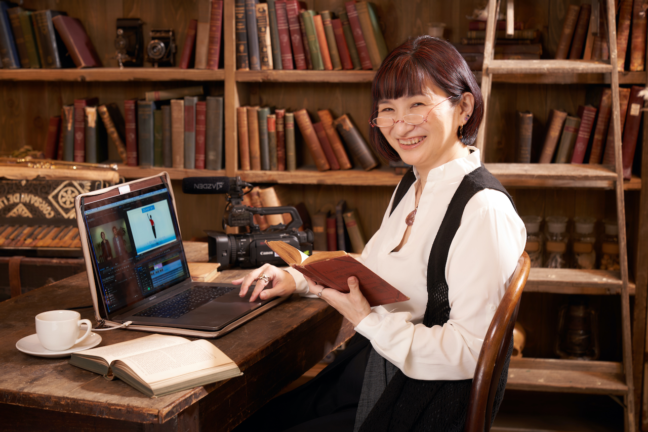古書の本棚の前でパソコンを開いて動画編集をしながら本を読み微笑む佐藤安南