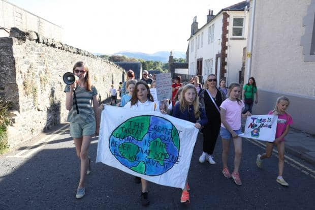 A group of around 20 schoolchildren marching up a street, one is holding a megaphone and two others are holding a sign which reads 'The greatest threat to our planet is the belief that someone else will save it'.