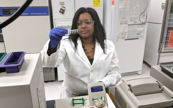 A woman in a lab coat (Samantha Maragh) looks at liquids in small vials.