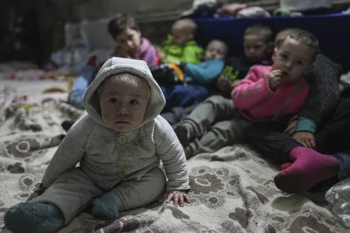 Toddlers sit on a blanket in a bomb shelter. 