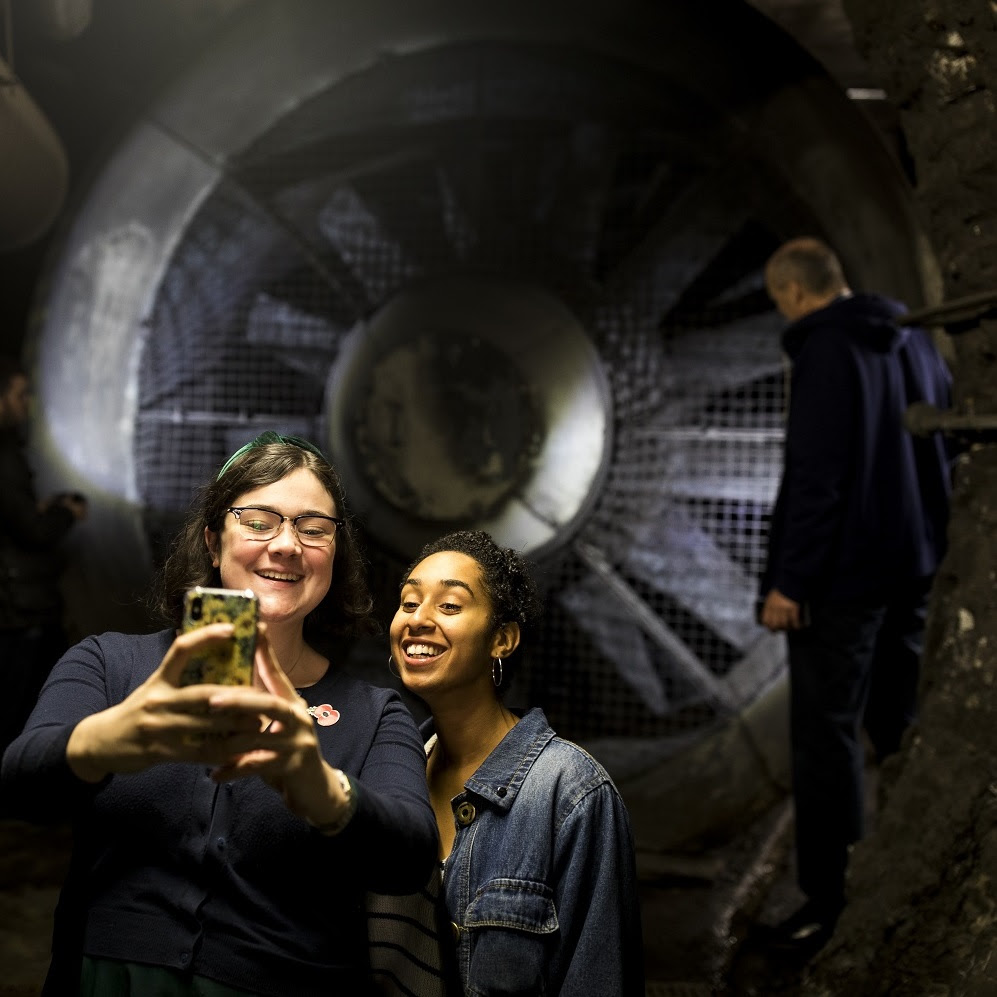Two people take a selfie in an industrial tunnel in front of a man looking at a huge fan