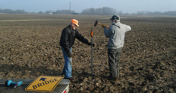 two men pound a post with an orange snowmobile trail marker into the ground
