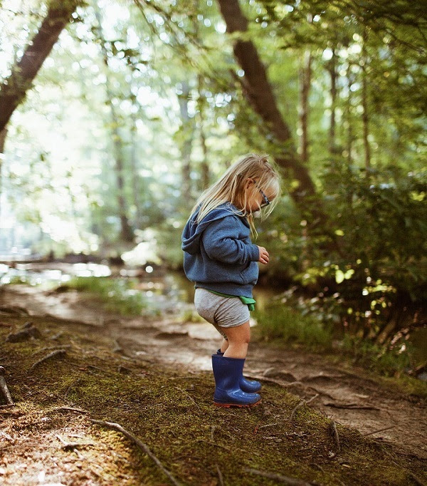a little blond-haired girl in navy blue rubber boots, dirt-scuffed shorts and a light blue jacket and glasses on stick-littered forest trail