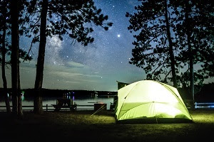 a yellow tent, glowing with inside light, nestled among shadowed trees next to water and a star-filled night sky