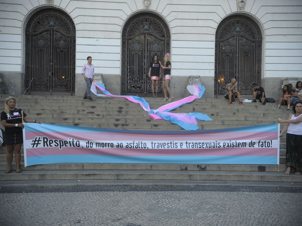 Duas mulheres seguram faixa comprida, com cinco listas, sendo a superior e a inferior azul calara, seguidas de duas faixas rodas, acima e abaixo de cada lista azul, e uma faixa branca ao centro, onde está escrito em letras pretas: "Respeito, do morro ao asfalto, travestis e transexuais existem de fato!" A faixa está na frente de uma escadaria de um prédio antigo, com três portas em arco ao fundo, onde algumas pessoas estão encostadas perto de outras panos (faixas ou bandeiras) nas mesmas cores, jogadas nos degraus.