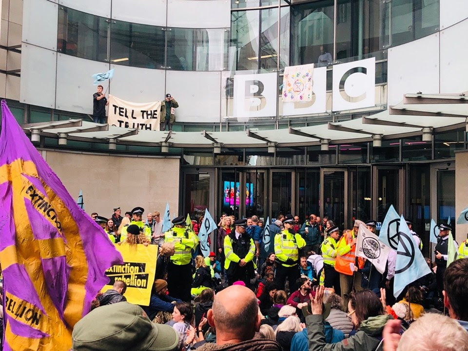 An XR demonstration in front of the BBC building in London. Some rebels are sat on the floor in front of the entrance, where a line of police officers stand behind them. Two (Scottish!) rebels are on the roof of the entrance doors with a sign that reads 'TELL THE TRUTH'.
