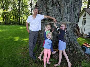 man and three young girls measuring base of large tree