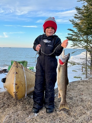 a young, smiling boy in winter gear and hat stands ashore in front of yellow canoe, holding a large silver fish from a chain