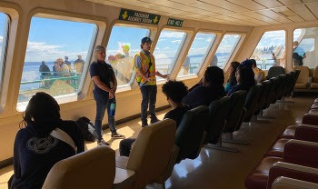 Two people speaking to several others that are seated inside the passenger cabin of a ferry