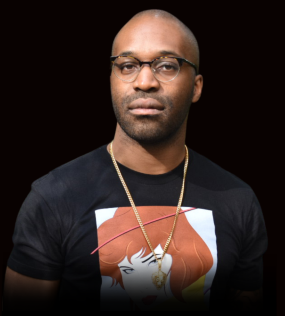 Tochi looks directly at the camera on an all black background. He is wearing glasses, a black t-shirt, and a gold necklace.