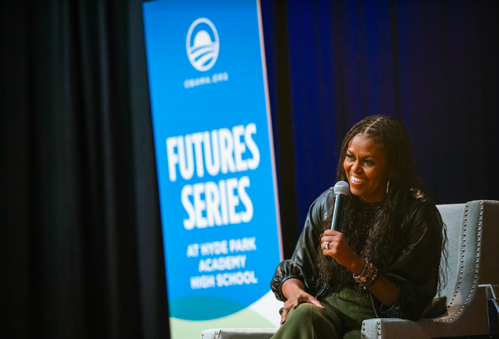 Michelle Obama smiles as she sits and holds a microphone at Hyde Park Academy High School in Chicago, IL. A sign in the background reads, “Futures Series at Hyde Park Academy High School.” 