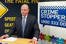 A police officer in a dark coloured suit and blue tie standing in front of road safety and crime stoppers signs.