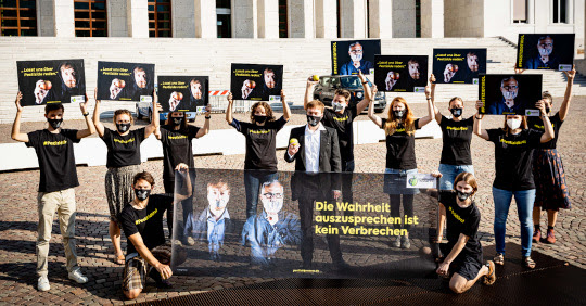Group of people stand outside building wearing black t shirts and holding placards with yellow writing