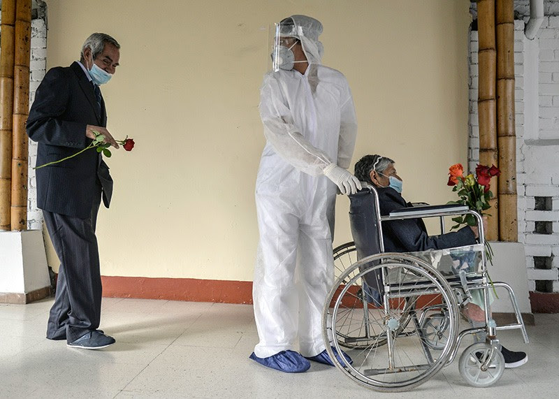 An elderly man holding a flower follows a worker pushing his wife in a wheelchair as she leaves intensive care.