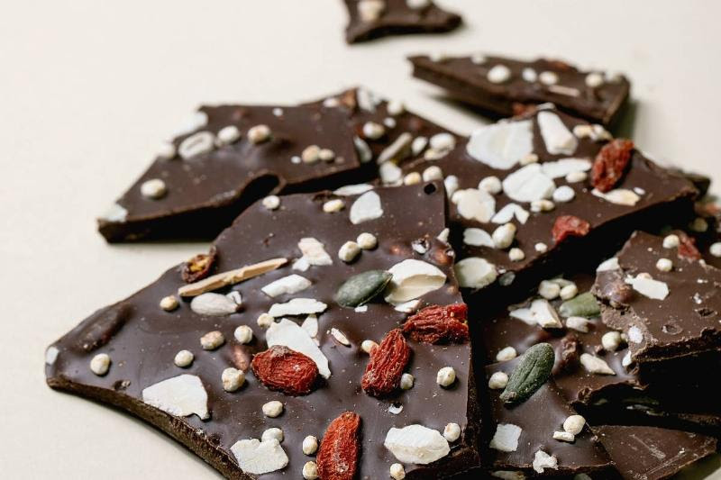 Handmade chopped dark chocolate with different superfood additives seeds and goji berries over beige background.