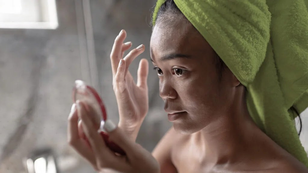 A woman applying serum to her face in a steamy bathroom, with her hair wrapped in a towel.