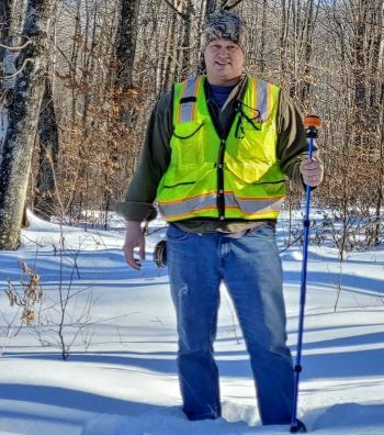 Forester Brock Van Oss stands in a snowy forest wearing a high visibility vest and a stocking cap. 
