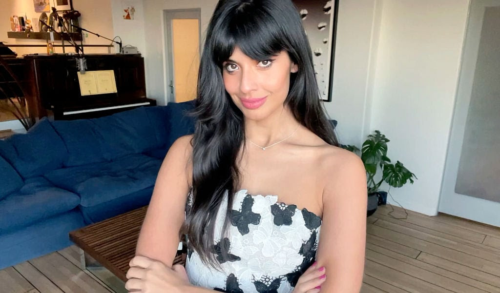 Jameela Jamil, a woman with long black hair and a white dress with black butterflies has her arms folded in front of her body and is smiling slightly.