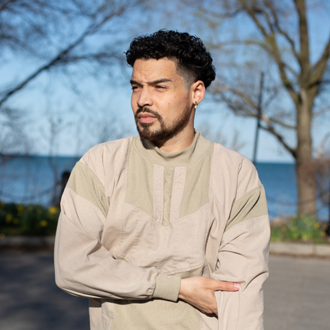 Light skinned Black femme with short, dark curly hair and beard in a sand colored sweatshirt posing in front of trees with Lake Michigan in the background.