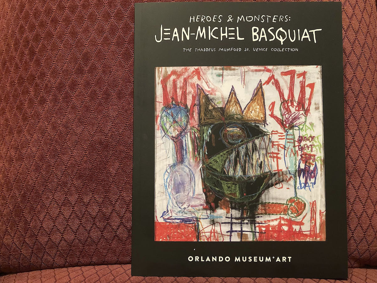 A book with the words 'Heroes & Monsters: Jean-Michel Basquiat' above a long-fingered creature with sharp teeth and a crown.