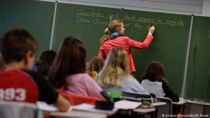 Quatro alunos sentados uns atrás dos outros em sala de aula na Alemanha. Ao fundo, professora escreve questão em inglês numa lousa verde