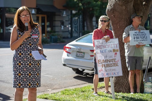 Kelly Wilkerson, organizer for Sister District CA-03, speaks at the rally. In the background, activists hold signs reading "The Senate minority is not the boss of us!! Eliminate the filibuster and restore democracy" and "We demand democracy 4 the people- no excuses"