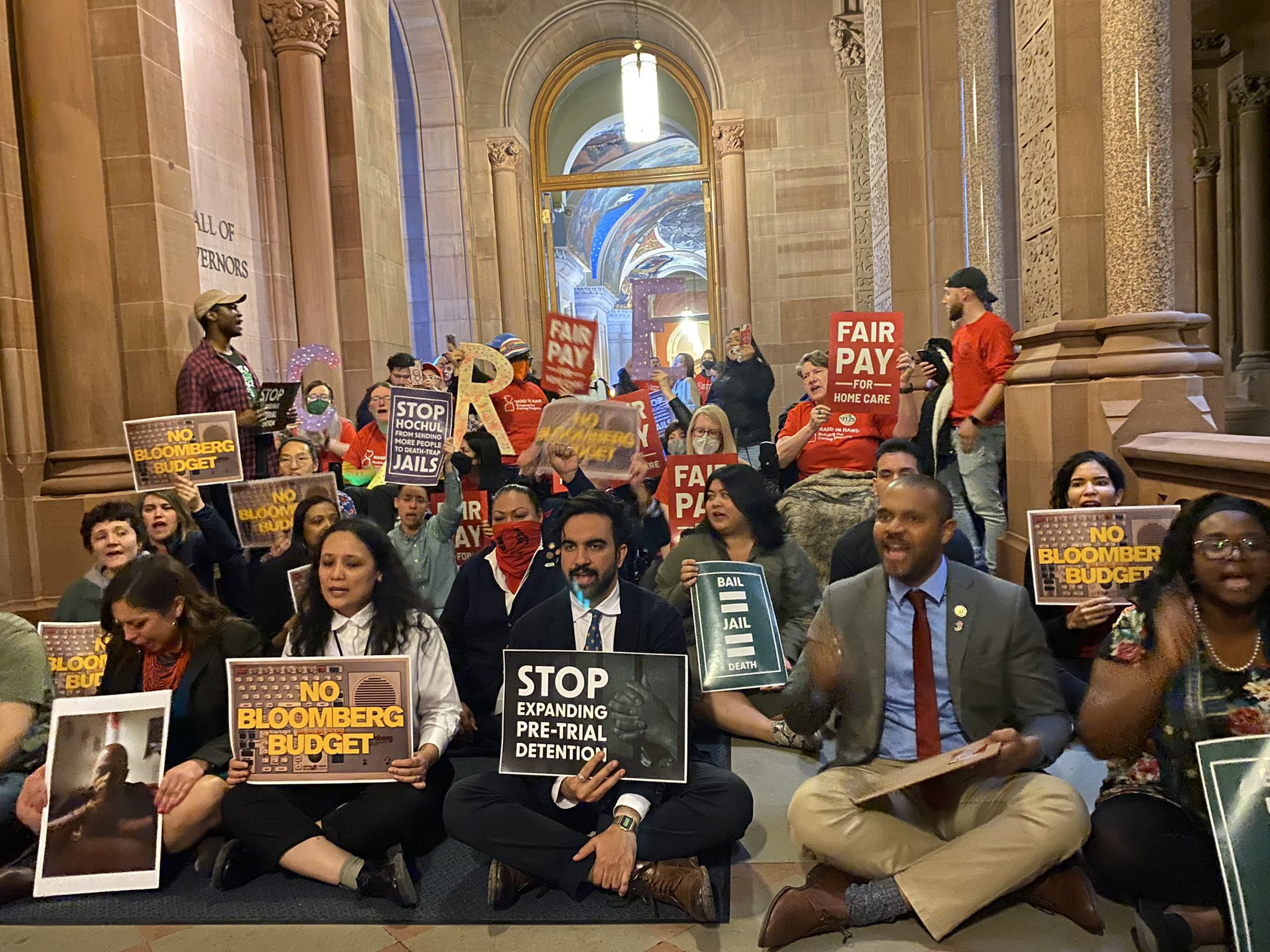 Photo of a sit-in at the State Capitol, with State Legislators sitting at the front, and JFREJ and Caring Majority members behind them.