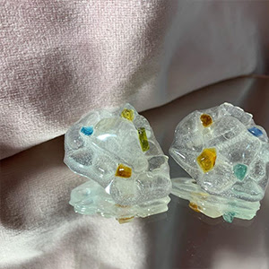 A photograph of a pair of stud earrings, they are transparent and textured with colourful small dots. 