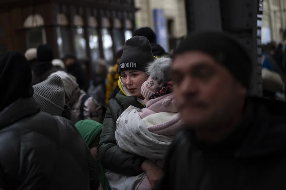 A woman holds her baby while surrounded by other people at a railway station.